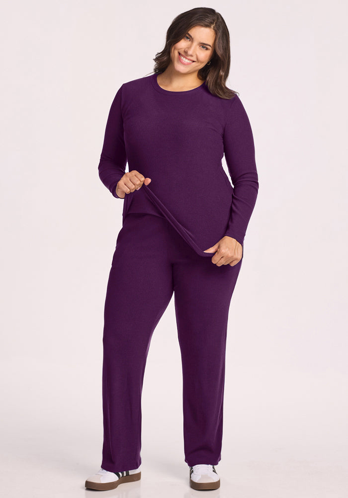A woman with dark hair smiles, wearing the Woolx Hadley Ribbed Crew in Majestic Purple. She pairs the moisture-wicking long-sleeve top with matching pants and white sneakers with black stripes, standing against a plain light background.