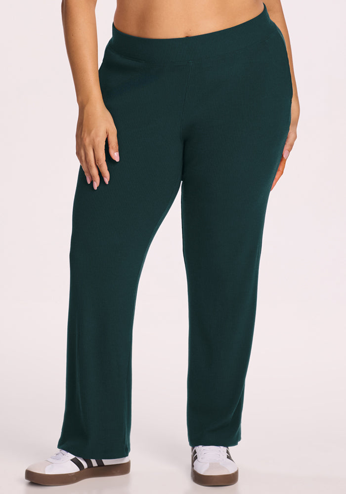 A person wearing Woolx Luca Ribbed Pants in Deep Timber and white sneakers with black stripes stands against a light background, showing only their lower torso and legs. 