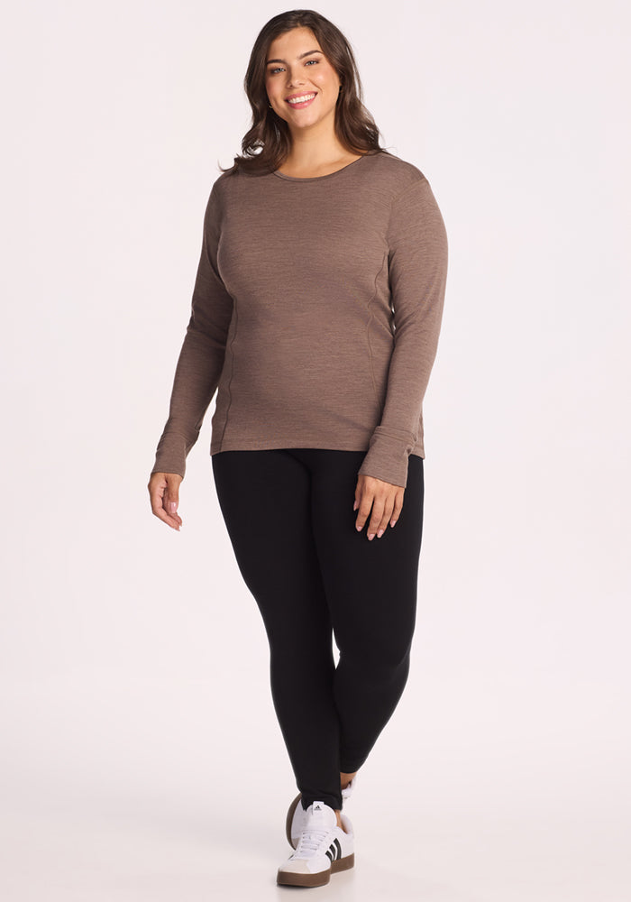 A woman with long brown hair is smiling and walking, wearing the Woolx Hannah Long Sleeve Top in Simply Taupe, black leggings, and white sneakers with black stripes against a plain light background.