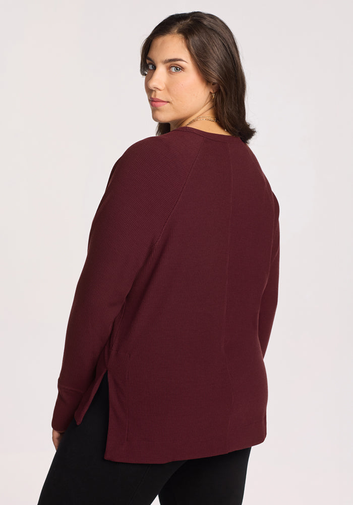 A woman with long brown hair, wearing the Woolx Ainsley Sweater in Toasted Garnet and black pants, stands facing away and looks over her shoulder at the camera against a plain light background.