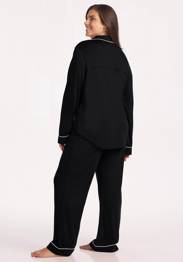A woman stands barefoot, looking over her shoulder and smiling in the Harper FeatherTouch® Pajama Set in black from Woolx, featuring white piping on the cuffs and hems, against a plain light background.