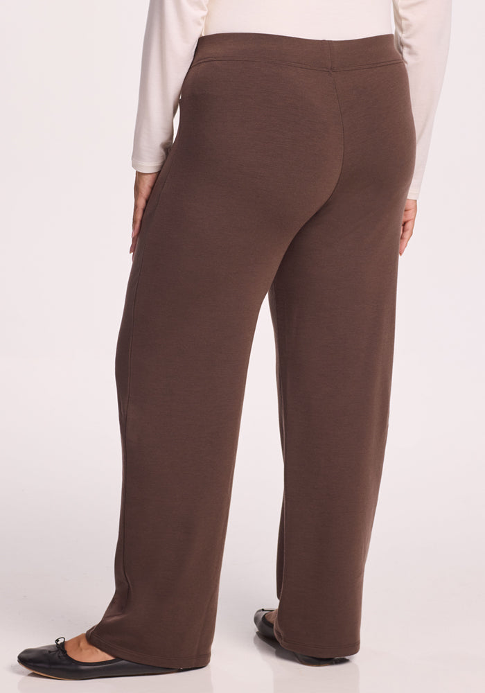 A person wearing Woolx Ellie Wide Leg Pants in Bracken and a long-sleeve white top stands facing away from the camera. The straight-leg pants are paired with black flats for a cozy, stylish look.