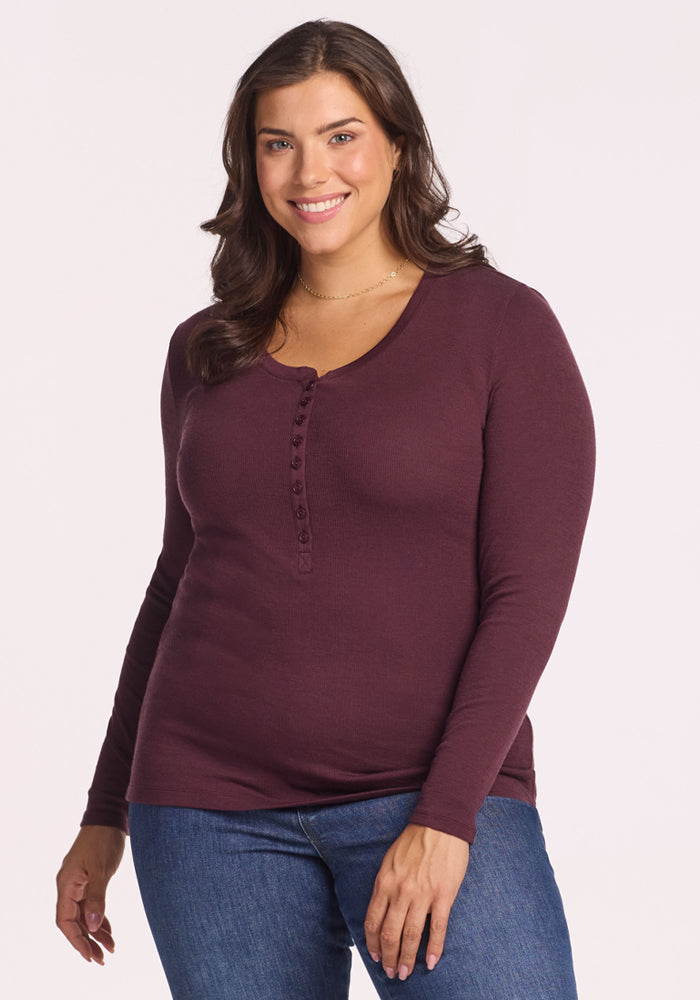 A woman with long brown hair, dressed in the Woolx Reese Ribbed Henley in Chocolate Fig and blue jeans, stands smiling against a plain light background. 