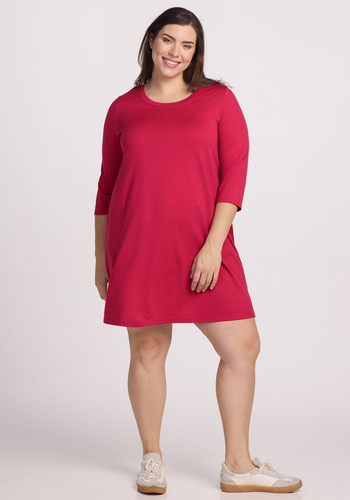 A woman with long brown hair smiles in the Woolx Rissa A Line Dress - Poppy Spark, a knee-length, bright pink Merino wool dress with three-quarter sleeves and white sneakers, set against a plain light background. 