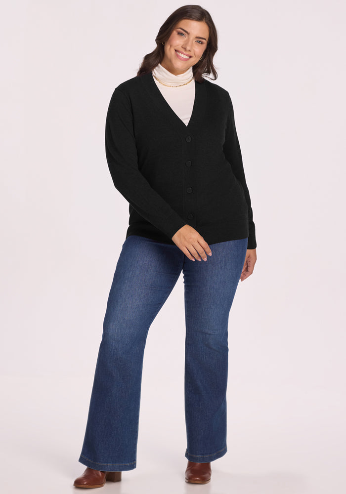 A woman with long brown hair smiles, wearing the Woolx Gia Sweater in black over a white turtleneck, blue jeans, and brown heeled boots. She stands with one knee slightly bent against a plain light background.