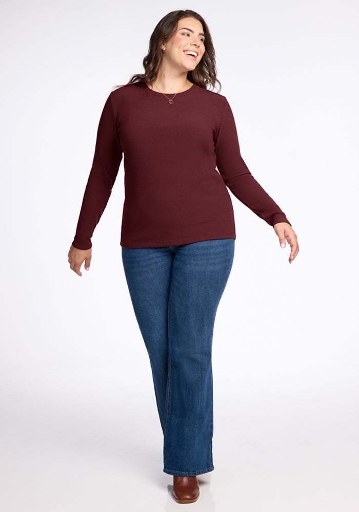 A woman with long brown hair, wearing the Woolx Hadley Ribbed Crew in Toasted Garnet, blue jeans, and brown shoes, smiles while walking forward—highlighting a stylish travel look against a plain white background.