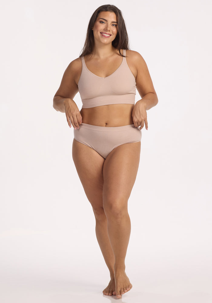 A woman with long brown hair stands barefoot, smiling and pointing down, wearing the Woolx Maeve Bikini in Sand against a plain white background.