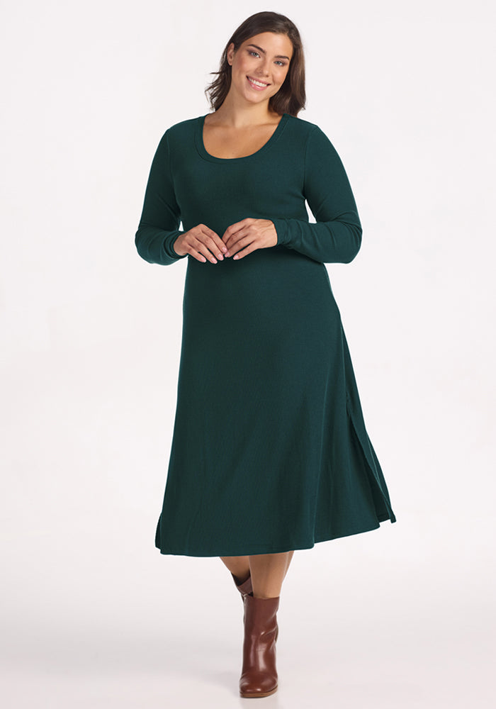 A woman in the Woolx Caroline Ribbed Twirl Dress in Deep Timber, paired with brown ankle boots, smiles against a plain white background. 