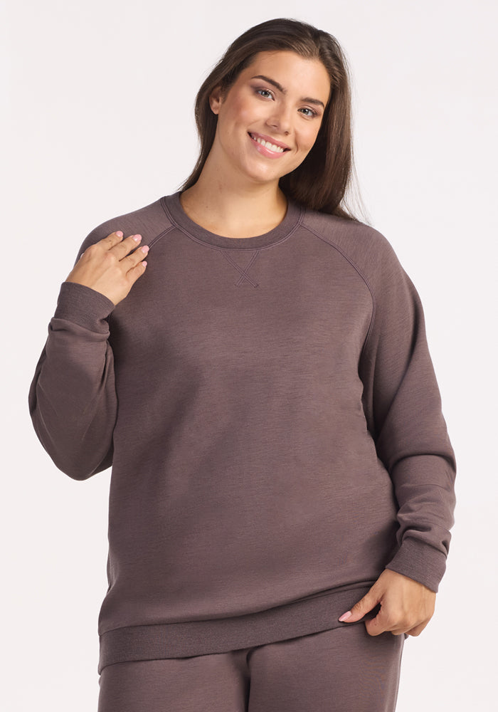 A woman with long brown hair is smiling and wearing the Woolx Bailey Crewneck Sweatshirt in Plum Truffle, paired with matching pants, as she stands against a plain, light background. 