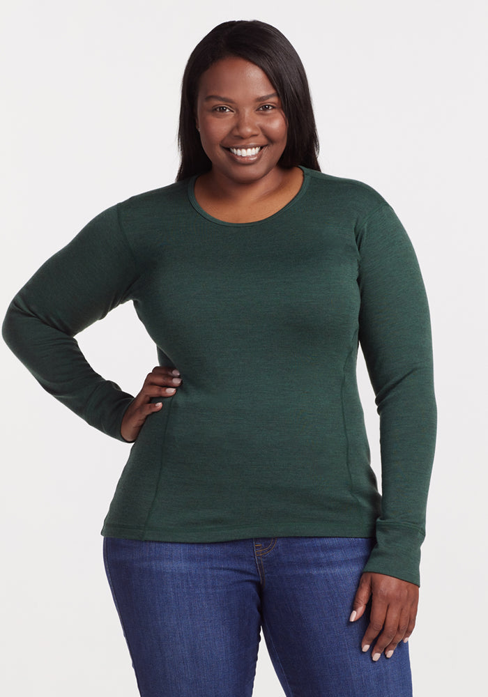 A woman smiles at the camera wearing the Woolx Hannah Long Sleeve Top in Forest with blue jeans, one hand on her hip, against a plain light background. 