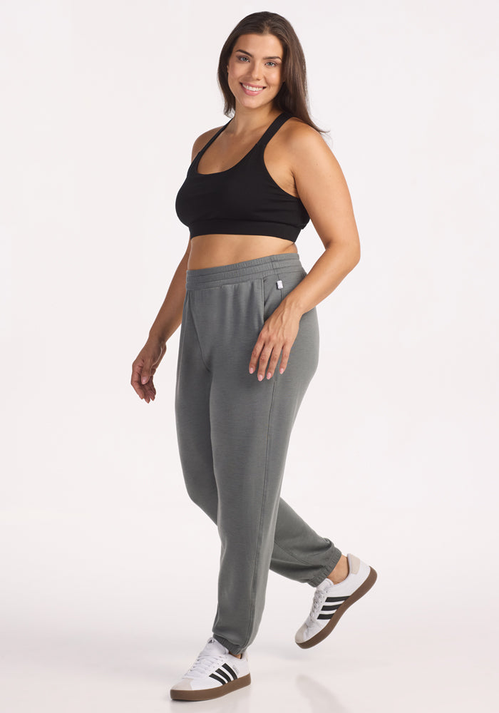 A woman with long brown hair smiles while standing sideways. She wears a black sports bra, Woolx Parker Sweatpants in Sedona Sage, and white Adidas sneakers with black stripes against a plain white background.