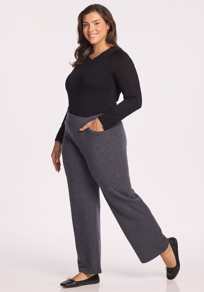 A woman stands smiling with one leg slightly raised, wearing a long-sleeve black top, Woolx Ellie Wide Leg Pants in Cinder Melange with pockets, and black flats against a plain white background.