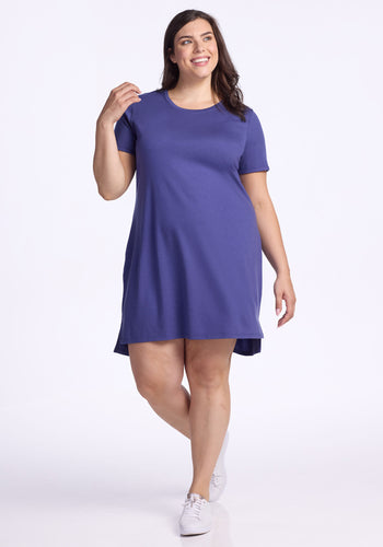 A smiling woman in the Woolx Lexie Dress & Cover-up - Ink Blue, a short-sleeved, knee-length dress made from breathable fabric, pairs it with white sneakers as she stands against a plain white background, one hand raised near her face. | Juliana is 5'10", wearing a size XL