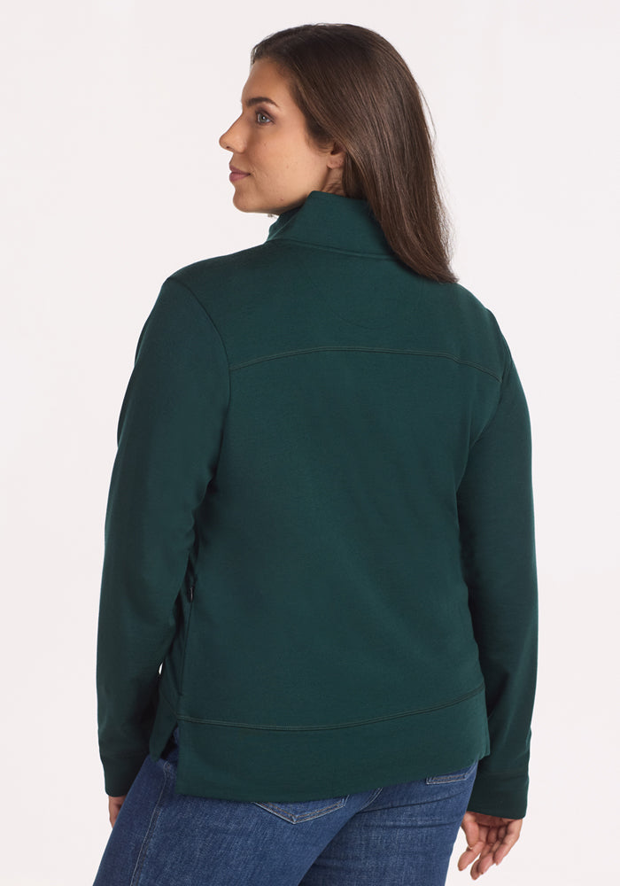 A woman with long brown hair wears the Woolx Annie 3 Button Pullover in Deep Timber and blue jeans, standing against a plain white background and looking over her shoulder.
