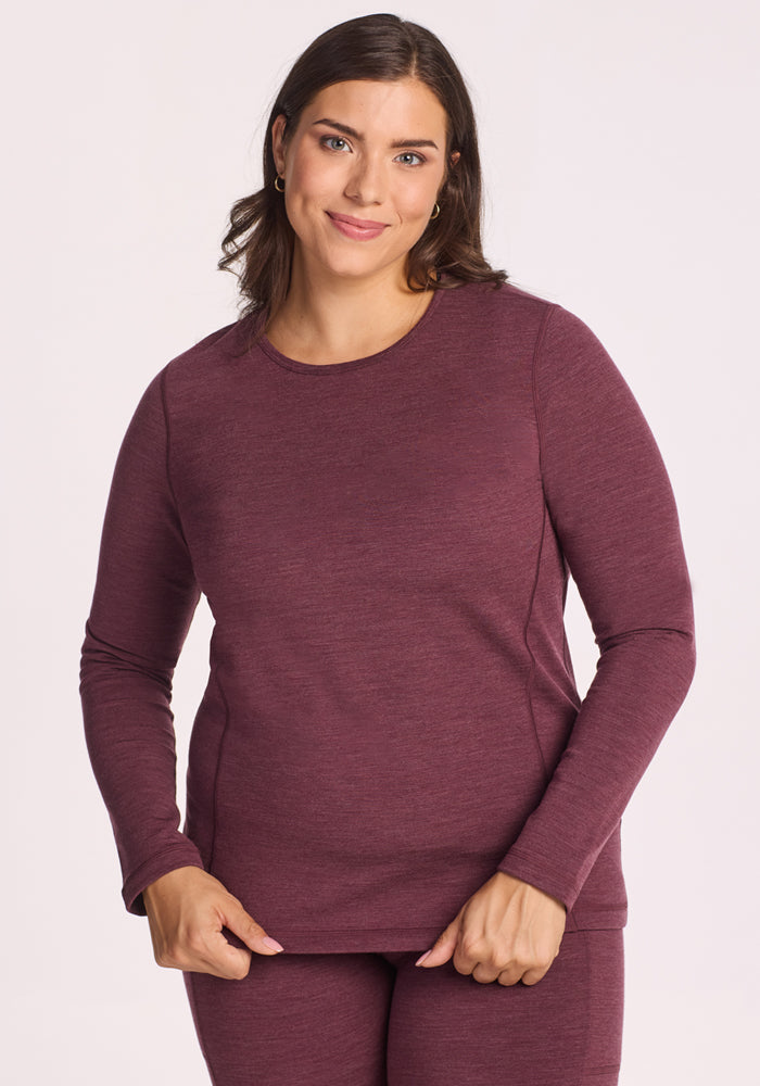 A woman with long brown hair wears the Woolx Elsa Long Sleeve in Grape Harvest Melange, an odor-resistant merino wool base layer, standing before a plain light background and smiling softly at the camera. 