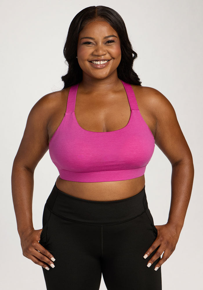 Smiling woman wears Woolx Livi Bra - Summer Sorbet and black leggings, standing with hands on hips against plain background. 