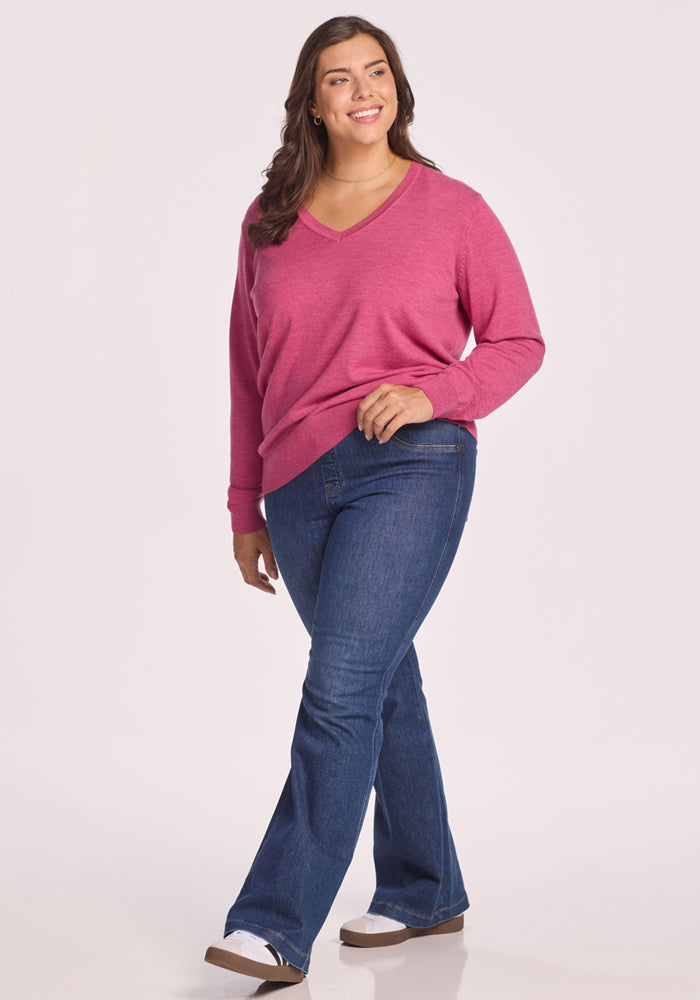 A woman with long brown hair wears the Woolx Francesca Sweater in Wild Salmon, styled with blue jeans and white sneakers, smiling against a plain white background.