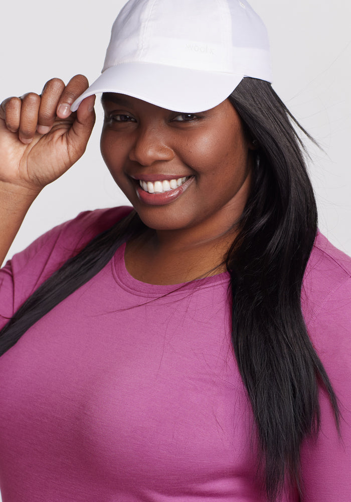 A woman in a Woolx Jenny 3/4 Sleeve Crew Neck - Purple Wine and a white baseball cap smiles, holding the brim of her cap against a light grey background.