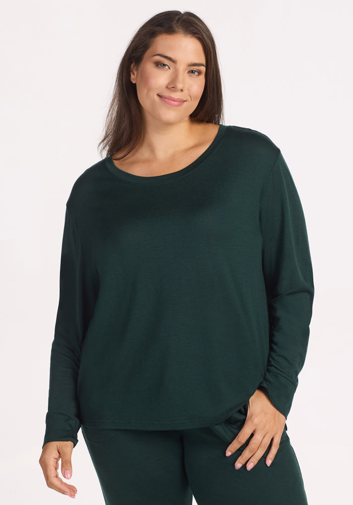 A woman with long brown hair wears the Woolx Ashlie Lounge Top in Deep Timber, standing against a plain white background and smiling softly with one hand in her pocket, highlighting the comfort of these moisture-wicking Merino wool pajamas. 
