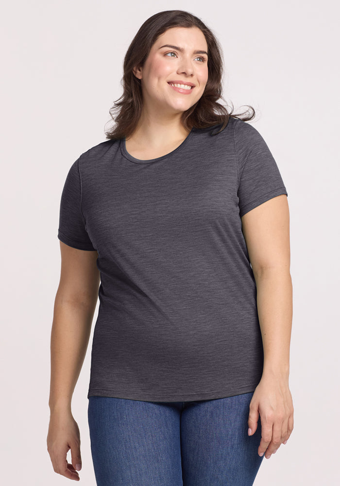 A woman with long brown hair smiles while wearing the Woolx Liza Crew Neck Tee in Cinder Melange and blue jeans. She stands against a plain, light background. 