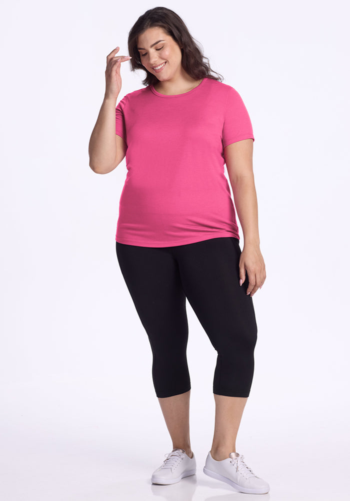 A woman with long brown hair smiles against a white background, wearing the Woolx Liza Crew Neck Tee in Raspberry Fizz, black capri leggings, and white sneakers. Her right hand touches her hair.