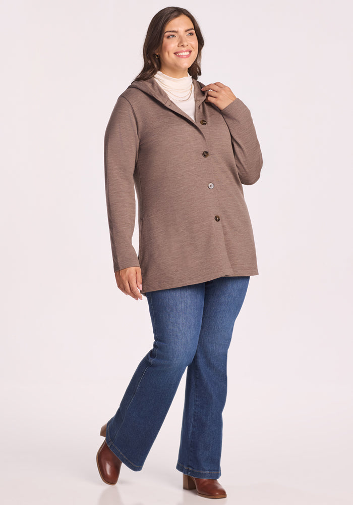 A woman smiles in the Woolx Edna Hooded Cardigan in Simply Taupe, styled with a white turtleneck, blue jeans, and brown ankle boots. She stands against a plain light background, perfectly dressed for chilly days.