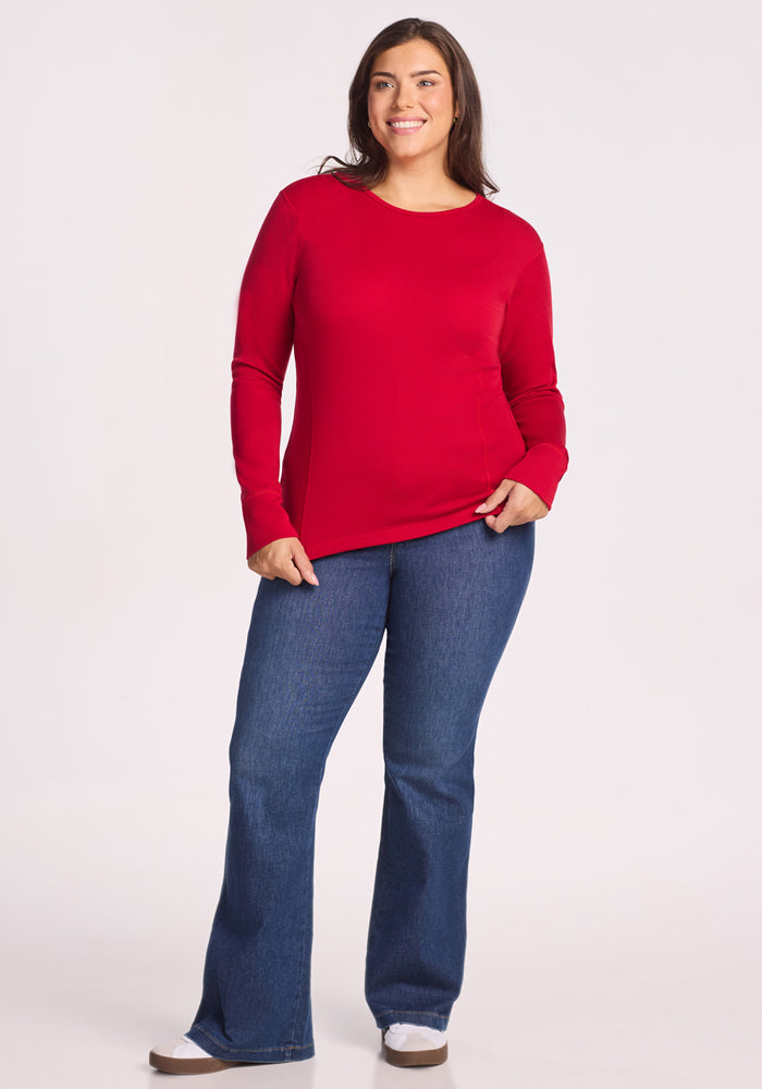 A woman smiles, wearing the Woolx Hannah Long Sleeve Top in Maraschino Cherry, paired with blue jeans and white sneakers. She has long brown hair and stands against a plain, light background.