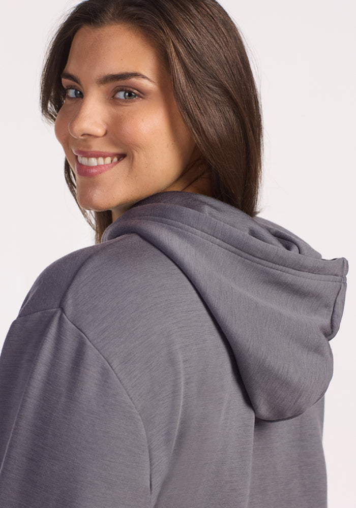 A woman with long brown hair wears the Woolx Avery Hoodie in Arctic Dusk and smiles over her shoulder at the camera, showing off its year-round comfort against a plain background.