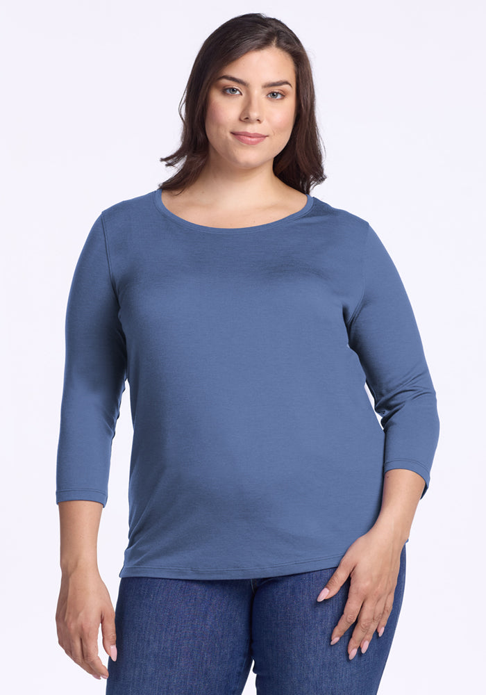 A woman with medium-length brown hair, wearing the Woolx Jenny 3/4 Sleeve Crew Neck in Skyline and blue jeans, stands against a plain light background, smiling softly at the camera. 