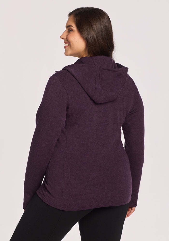 A woman with long brown hair smiles while wearing the Woolx Zoey Zip Up Hoodie in Deep Plum and black pants, standing with her back slightly turned to the camera against a plain light background.