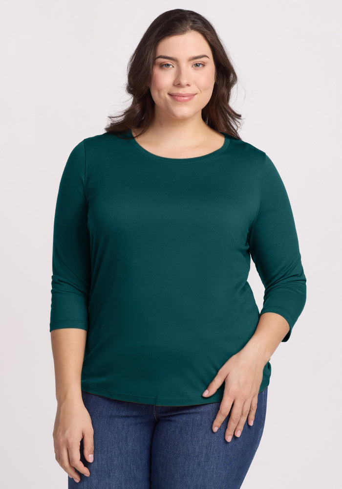 A woman with long brown hair, wearing the Woolx Jenny 3/4 Sleeve Crew Neck in Riverbank Green and blue jeans, stands facing the camera and smiles gently against a plain light background. 