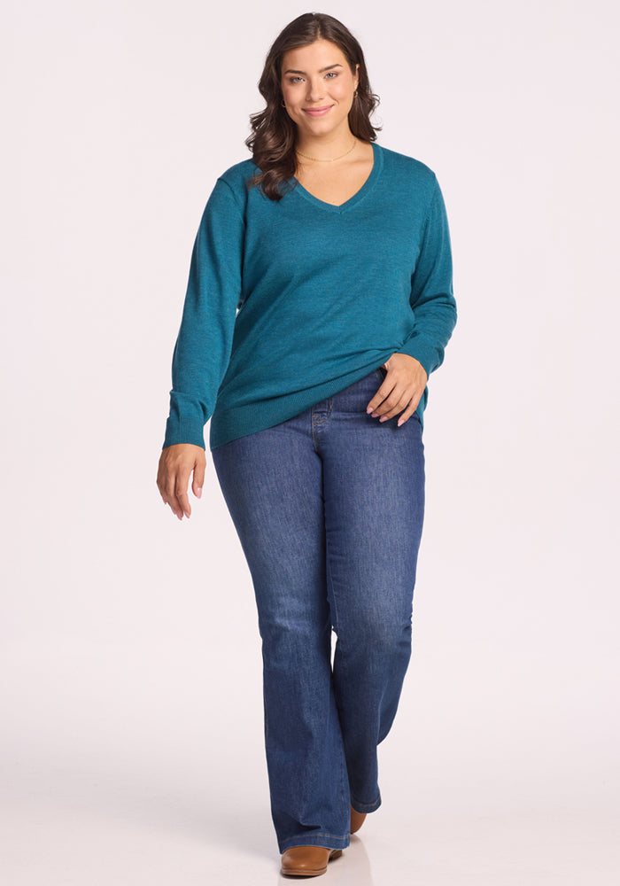 A woman with wavy brown hair smiles and walks forward in a Woolx Francesca Sweater in Harbor Teal, blue bootcut jeans, and brown shoes against a plain white background.