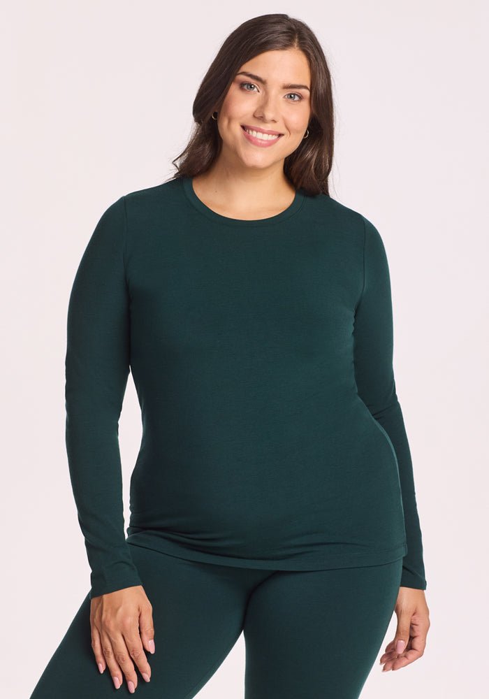 A woman with long brown hair smiles at the camera, wearing the Woolx Sadie Baselayer Top in Deep Timber, made from Duralite merino fabric, and matching pants against a light background. 