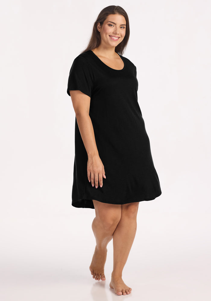 A woman with long brown hair, barefoot and smiling, stands on a light background wearing the Woolx Desi Nightgown in black, which is short-sleeved, knee-length, and made of Merino wool. 