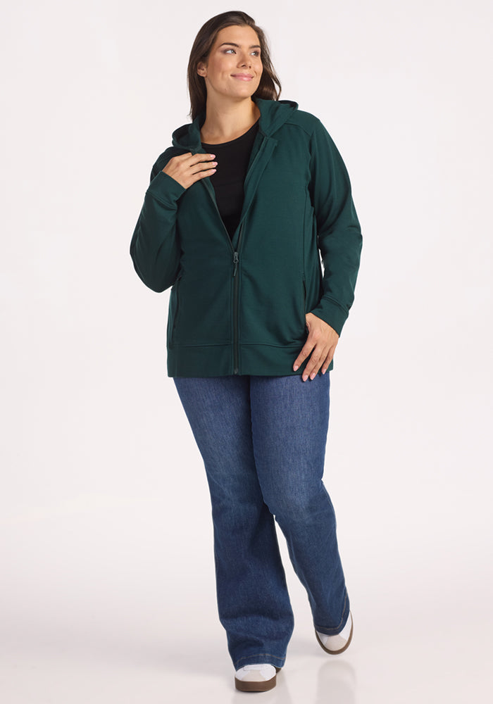 A woman smiles in the Woolx Darby Zip Up Hoodie in Deep Timber, layered over a black top and blue jeans. She touches her hood, looks to the side, and wears white sneakers against a plain white background.
