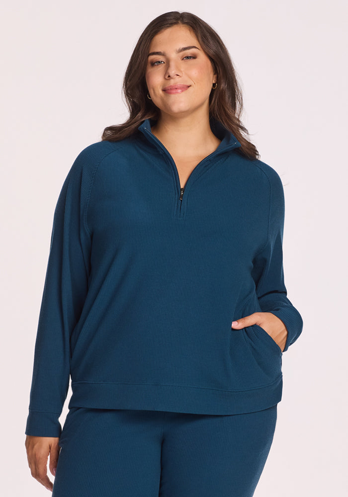 A woman with long brown hair smiles, wearing the Woolx Quinn Ribbed 1/4 Zip in Majolica Blue with matching pants, one hand in her pocket, against a plain light background. 