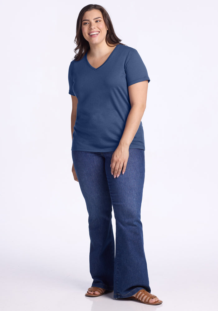 A woman with long brown hair, wearing the Woolx Junie V Neck in Starry Night, blue jeans, and brown sandals, stands and smiles against a plain white background.