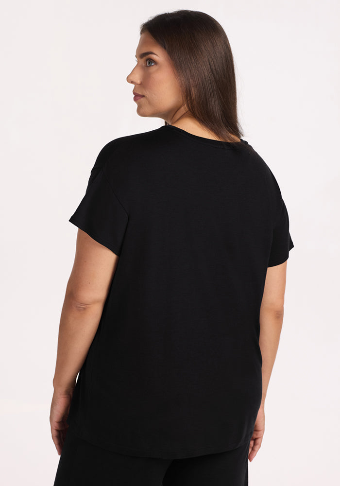 A woman with straight brown hair stands facing away, wearing the Woolx Billie Lounge Tee in black and matching pants—ideal as cooling sleepwear—against a plain light background.