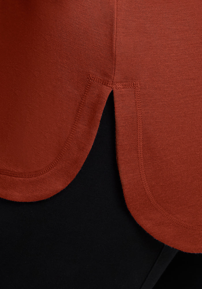 Close-up of the Blair Tunic - Maple Leaf by Woolx, showing the side seam with a curved hem and small side slit. The rust-colored Merino wool tunic is styled over black pants.