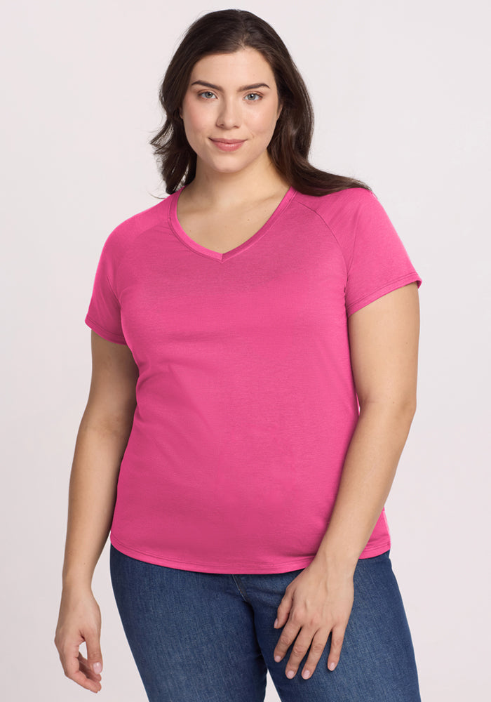 A woman with long brown hair wears the Woolx Mia V Neck - Raspberry Fizz Merino wool T-shirt and blue jeans, standing against a light background and looking at the camera with a slight smile. 