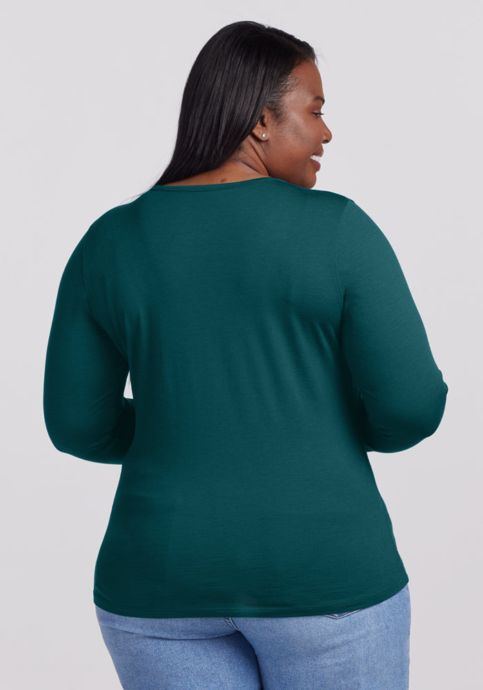 A woman with long black hair is seen from the back, wearing the Woolx Remi Long Sleeve T-Shirt in Riverbank Green and light blue jeans, set against a plain light background.