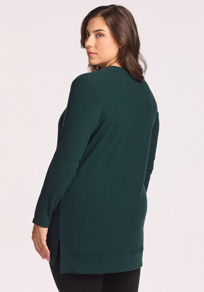 A woman with medium-length brown hair wears the Woolx Evie Ribbed Tunic in Deep Timber, a cozy long-sleeve sweater with side slits. She stands sideways, looking over her shoulder against a light background.