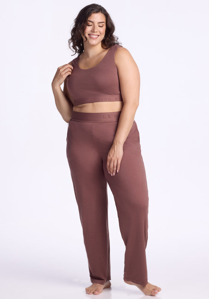 A smiling woman with wavy brown hair wears the Woolx Aubrey Lounge Bra in Nutmeg, standing barefoot against a plain white background.