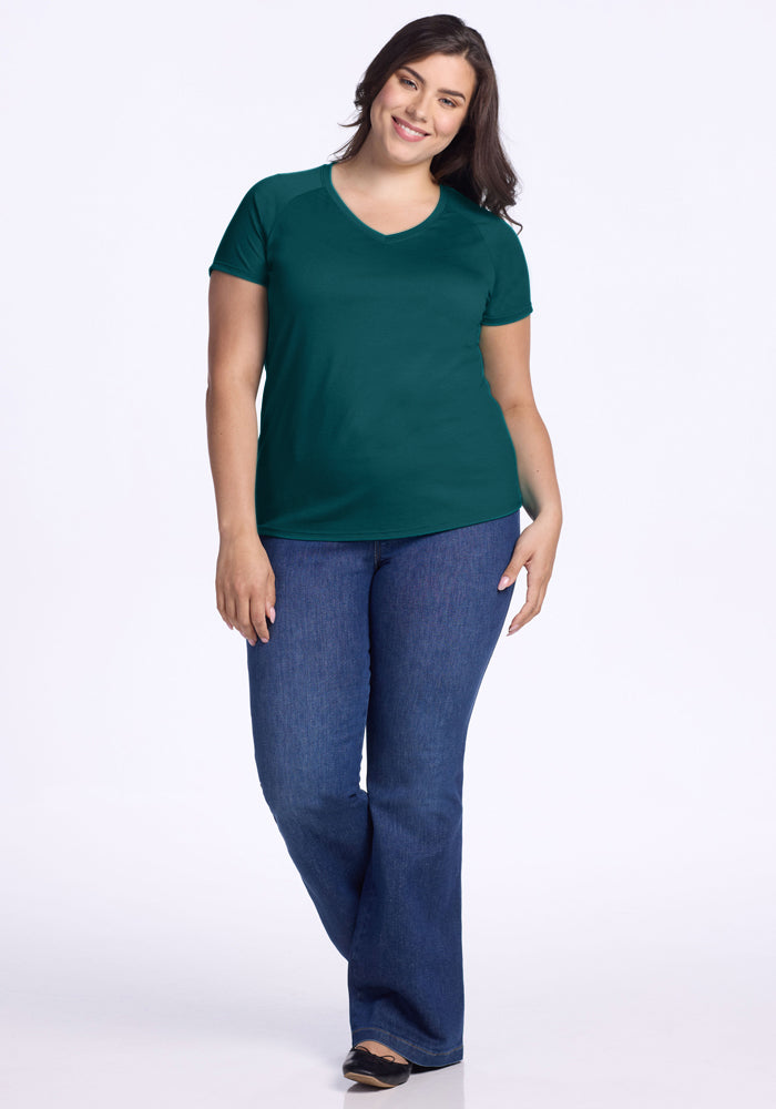 A woman with long dark hair stands smiling against a white background, wearing the Woolx Mia V Neck in Riverbank Green, a lightweight odor-resistant Merino wool t-shirt, along with blue jeans and black shoes.