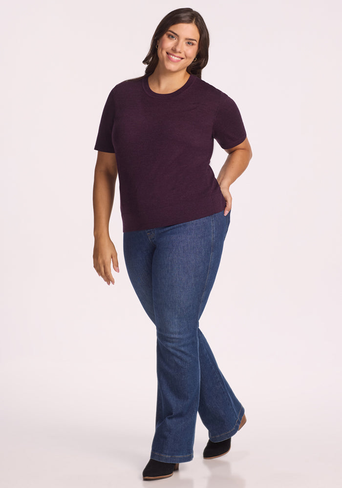 A woman with shoulder-length brown hair smiles while standing against a white background, wearing the Woolx Anna Crew Sweater in Deep Plum, paired with blue jeans and black shoes. Her left hand is on her hip, and her right arm rests at her side.