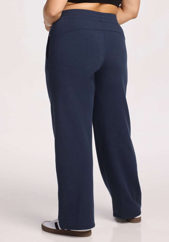 A person is seen from behind on a white background, wearing Woolx Brynn Pants in Deep Navy, a black top, and white slide sandals. The image highlights the relaxed fit of the heavyweight merino travel pants.