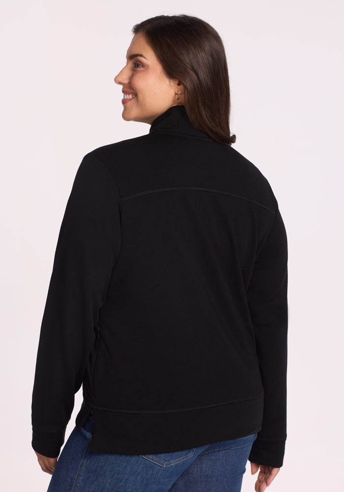 A woman with long brown hair models the Woolx Annie 3 Button Pullover in black, paired with blue jeans, smiling and looking over her shoulder to showcase the back against a light background.