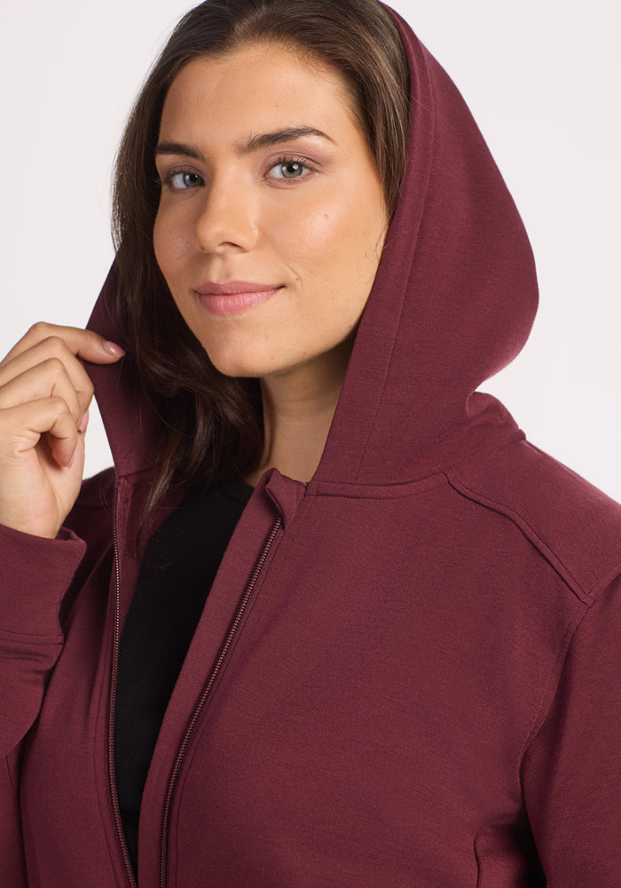 A woman smiles softly at the camera, holding the hood of the Woolx Darby Zip Up Hoodie in Toasted Garnet, highlighting its cozy DuraLite fabric against a plain background.