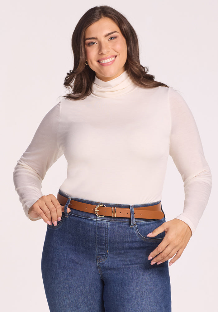 A smiling woman with long brown hair wears the Woolx Kennedy Bodysuit in Buttercream, paired with blue jeans and a brown belt. She stands against a plain light background, hands near her waist, highlighting this versatile layering essential. 