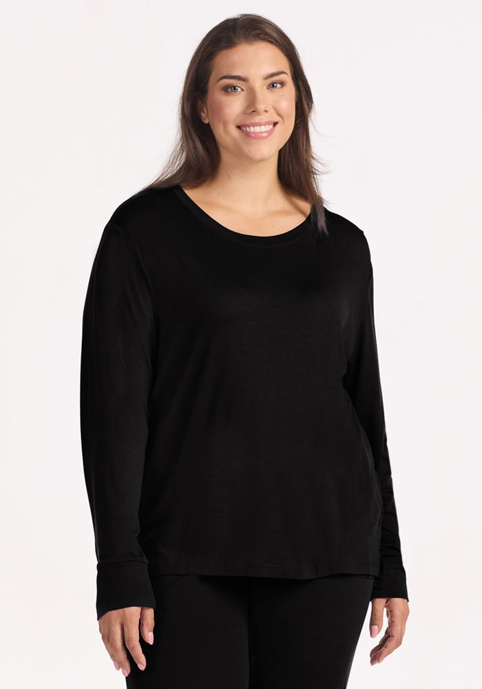 A woman with long brown hair, wearing the Woolx Ashlie Lounge Top in black, stands smiling against a plain white background. 